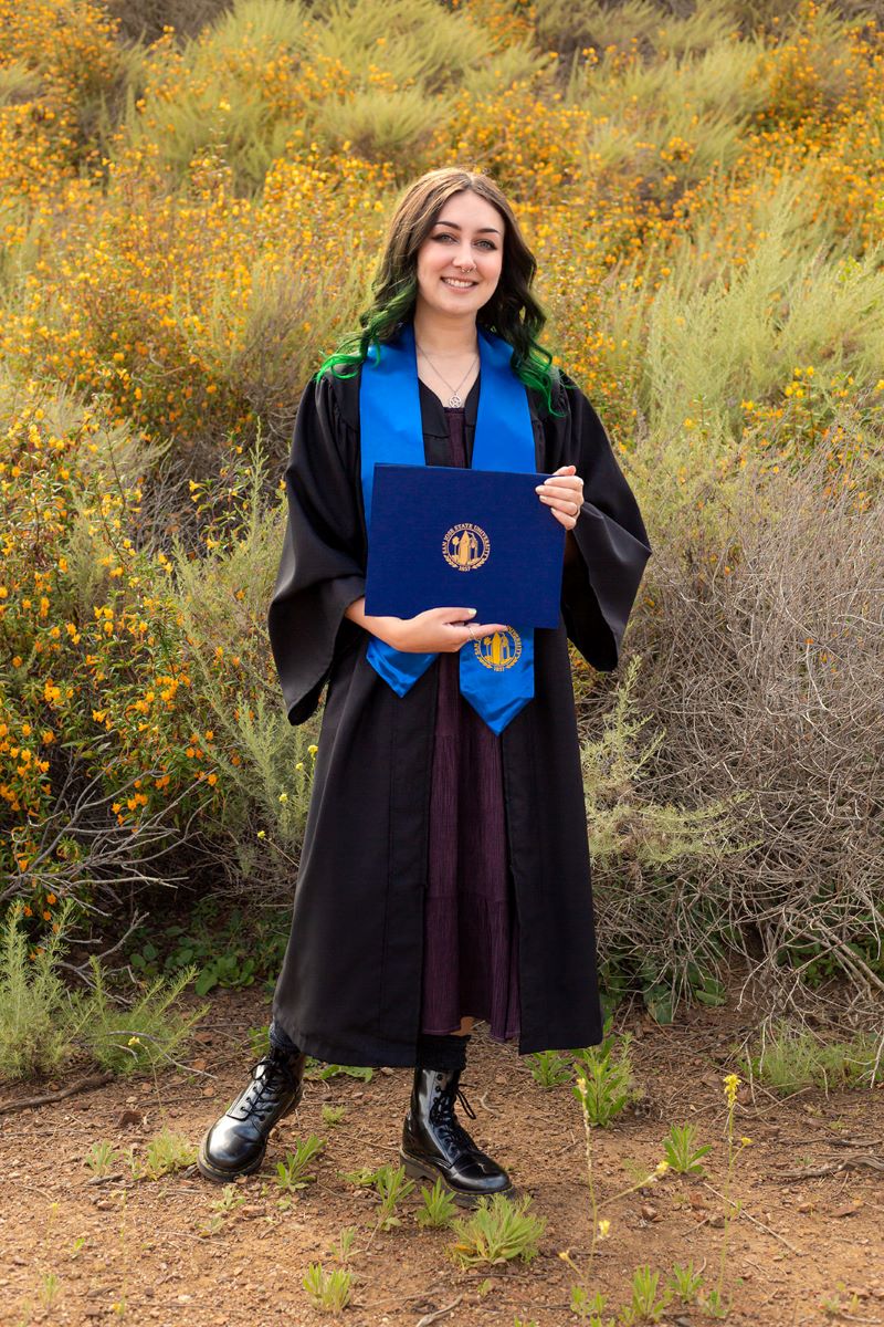 A person holding a navy blue diploma and wearing a blue stole and a black graduation gown and black boots