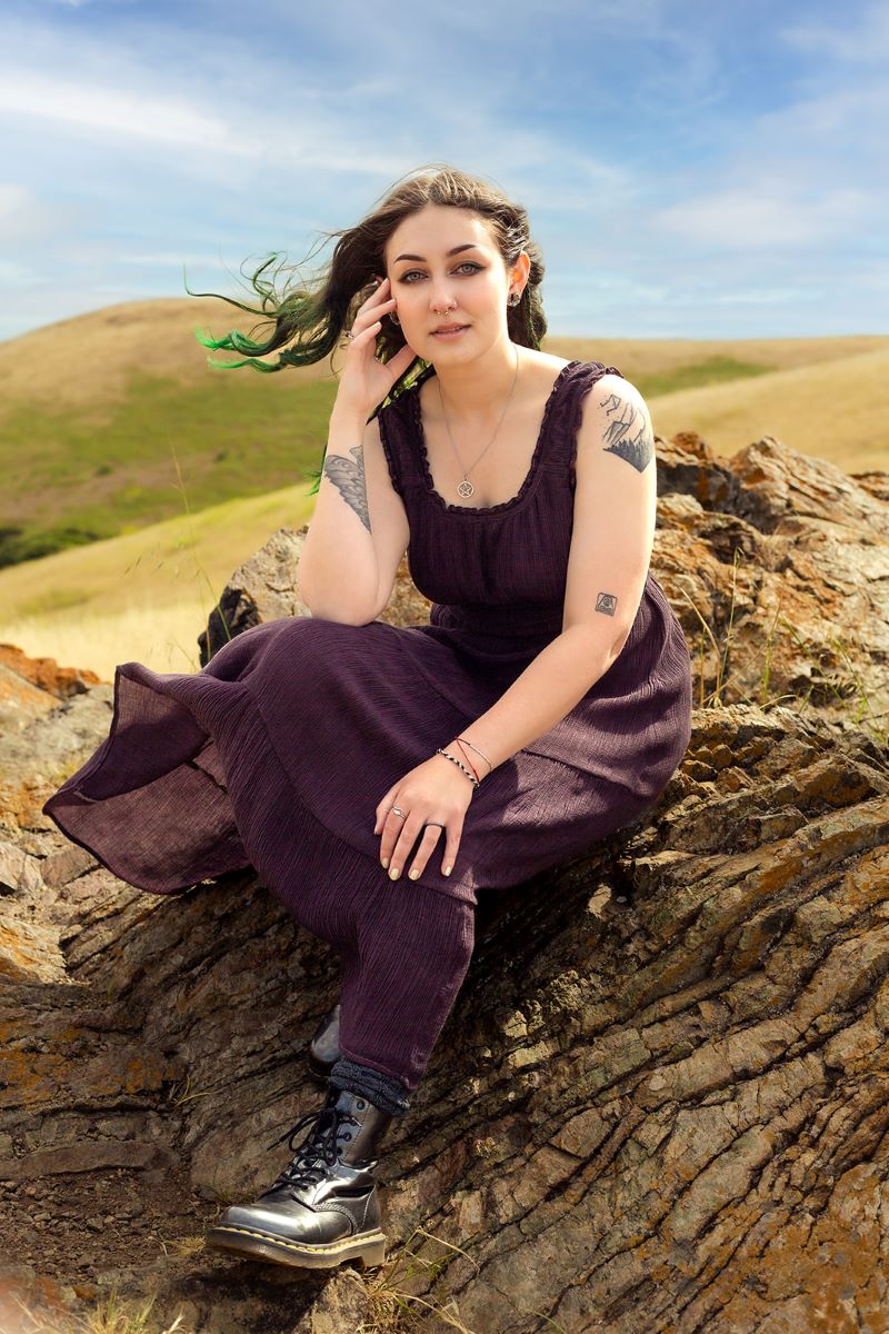 A person sitting on a rock. They are wearing a purple dress and black boots and they have tattoos. 