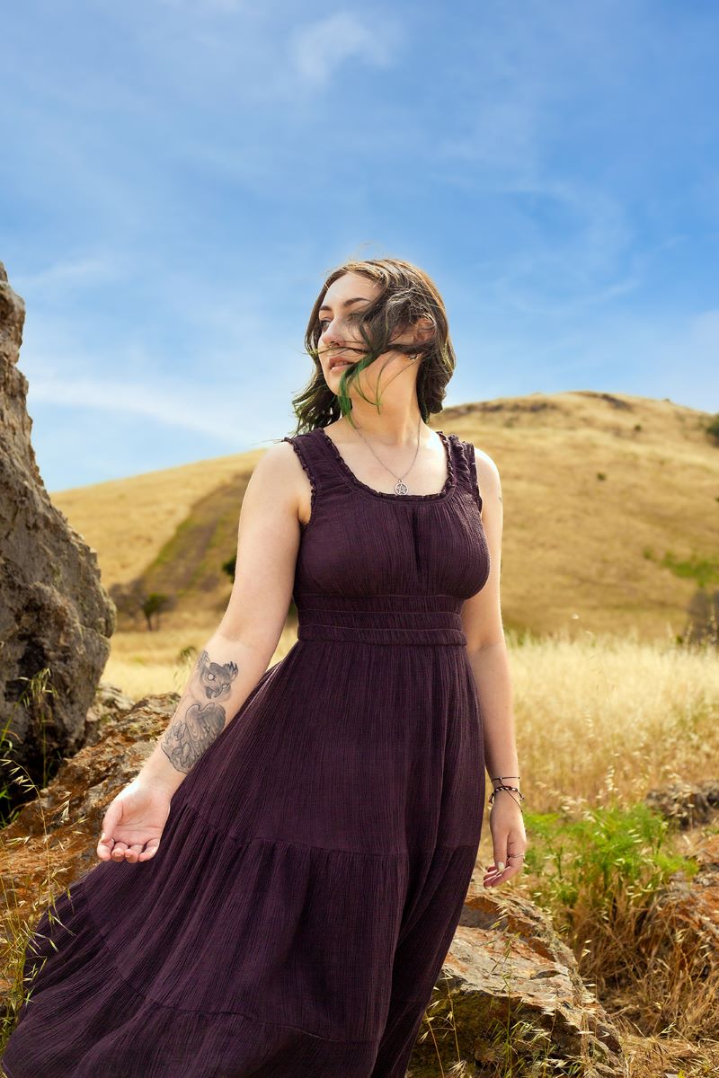 A person standing next to a rock in front of a hill looking off to the side. They are wearing a purple dress and have tattoos. 