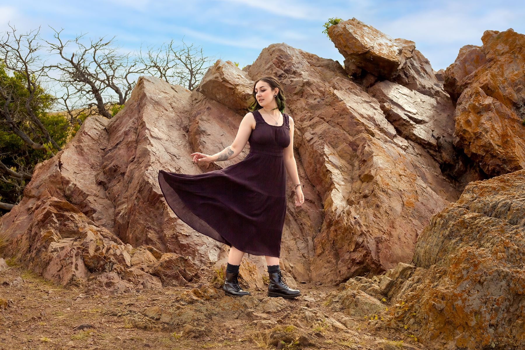 A person wearing a purple dress standing in front of brown rocks they are flipping their brown dress to the side. They are wearing black bloots and also have tattoos. 