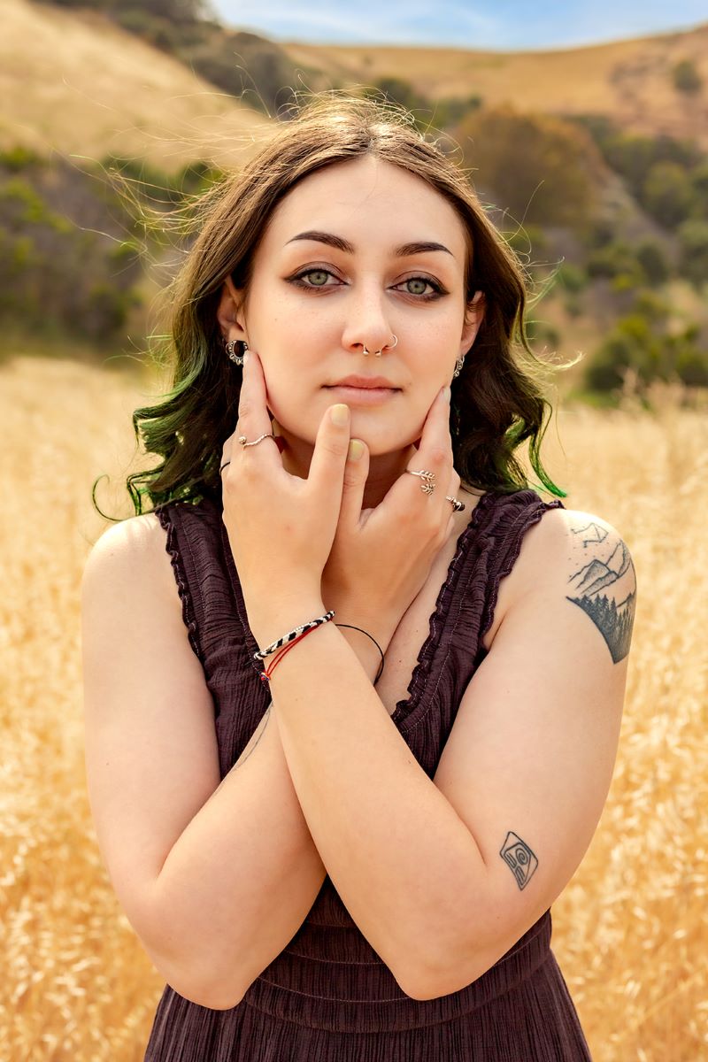 A person gently holding their face with their arms crossed. They have highlights of green in their hair and tattoos. 