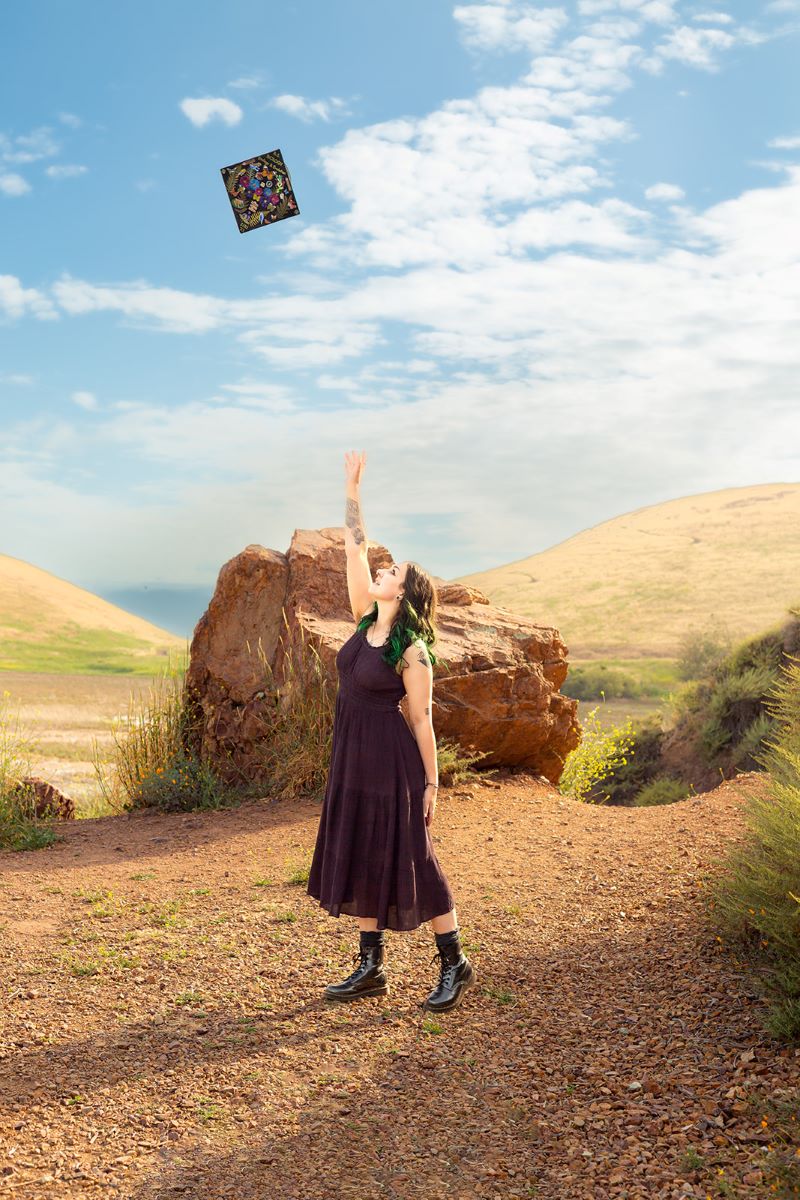 A person tossing their graduation cap into the air underneath a blue sky with clouds and behind them is a large rock. They are wearing a purple dress and have on black boots. 