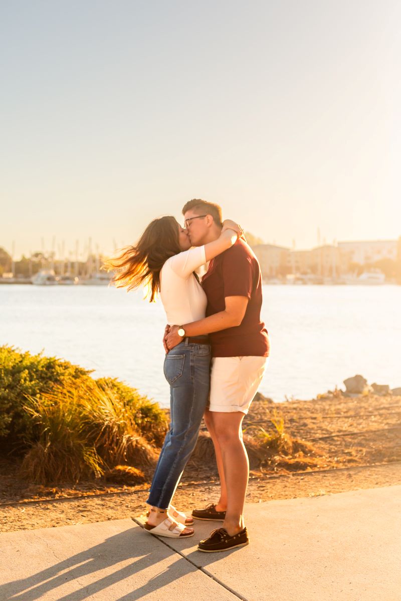 Couple kissing at Jack London Square in front of the marina one is wearing a white shirt and blue jeans and the other is wearing a red shirt and white shorts 