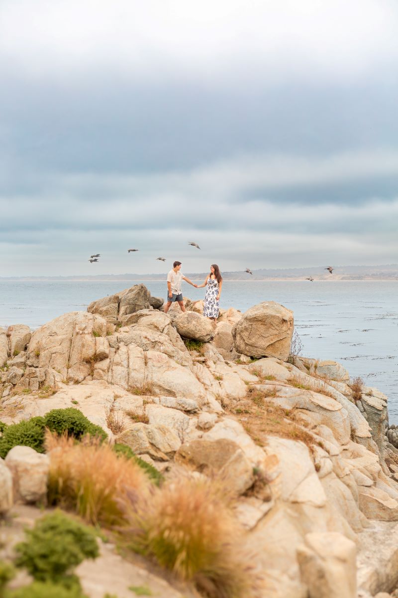 Couple holding hands on the rocks while seagulls fly behind them and the ocean is also behind them they are both looking at each other 