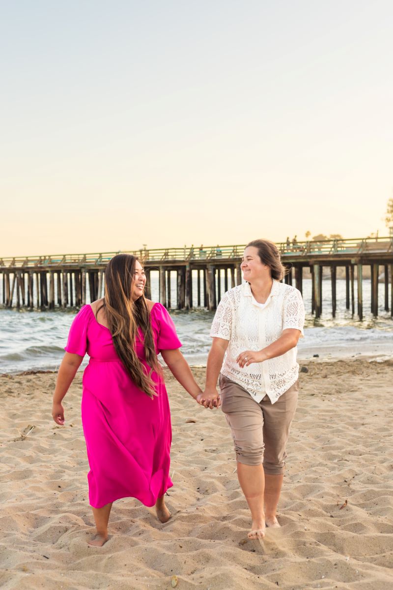 Couple walking hand in hand in the sand together one is wearing a neon pink dress and the other is wearing a white shirt and brown pants and both are looking at each other and laughing 