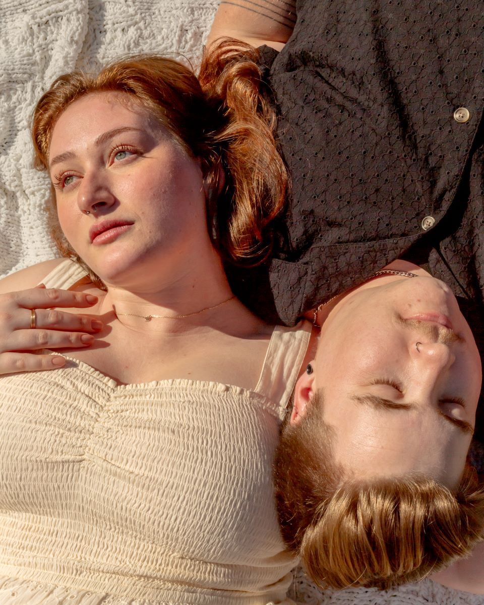 couple laying down on a blanket one is looking off to  the side and has a gold ring and necklace and a yellow dress and red hair and the other is wearing a black shirt and has a nose ring and earring 