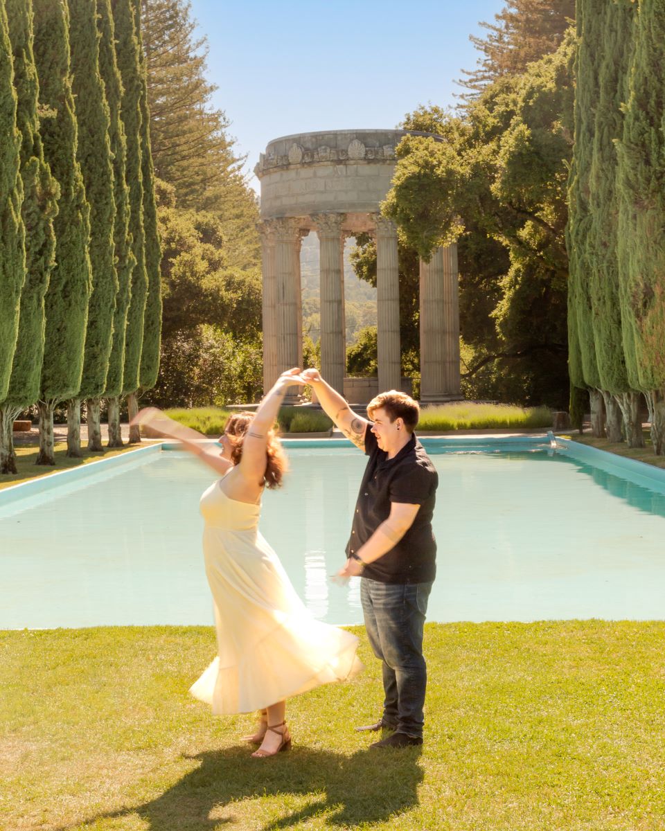 Couple at Pulgas Water Temple behind them is a reflecting pool and trees one persona is spinning the other who is wearing a yellow dress the other one is wearing jeans and a black shirt 