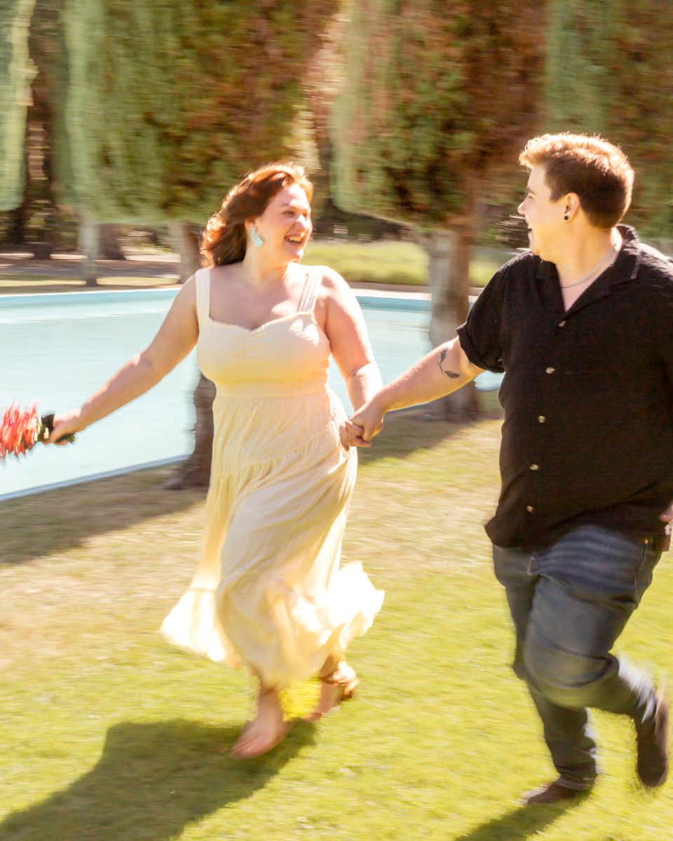 couple running in the grass next to the Pulgas Water Temple one is wearing a yellow dress, blue earrings, and holding flowers in one hand the other has on jeans and a black shirt 