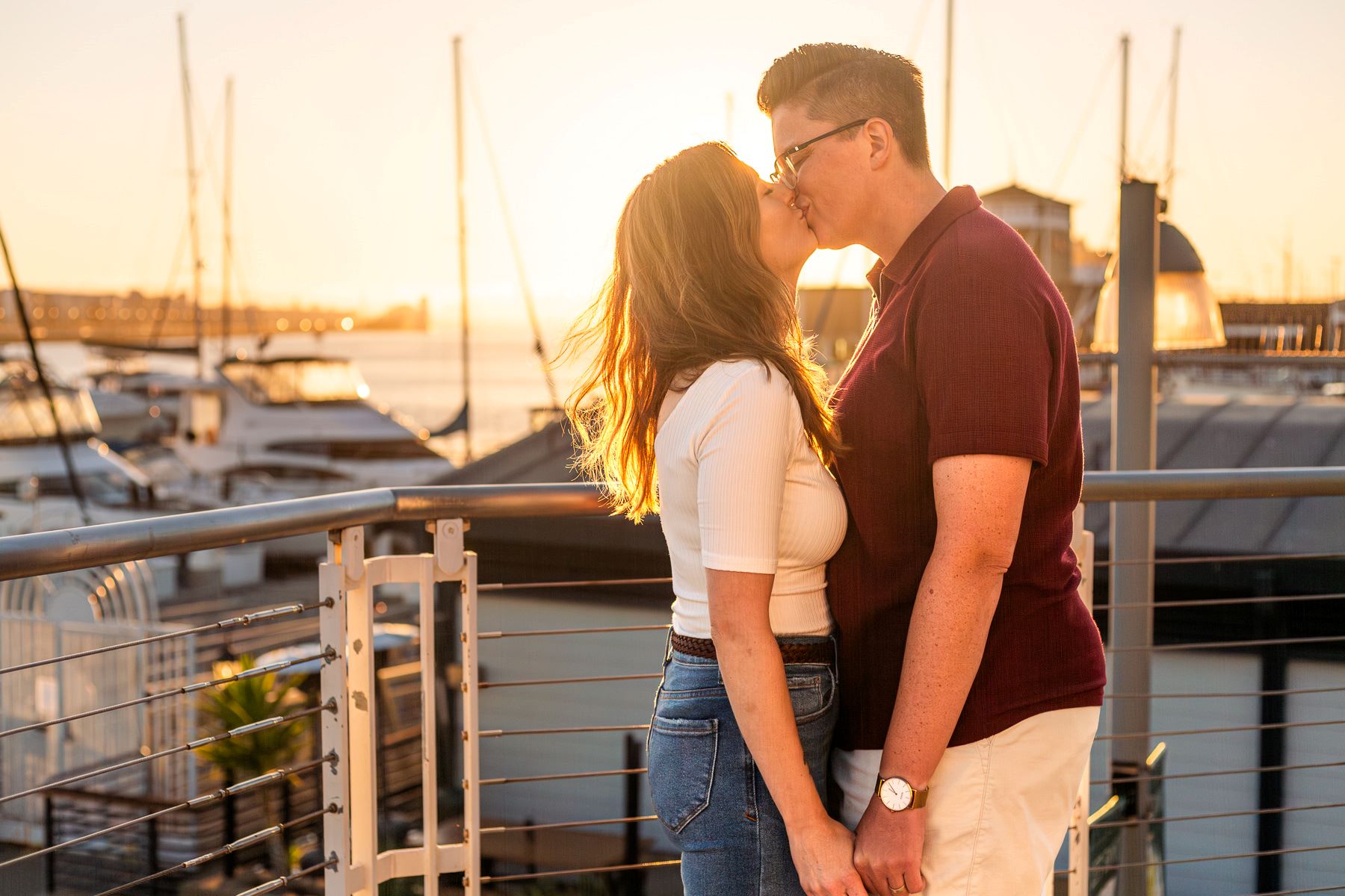 couple kissing each other at Jack London Square while the sun sets behind them one is wearing a white shirt and blue jeans and the other is wearing a maroon shirt and shorts