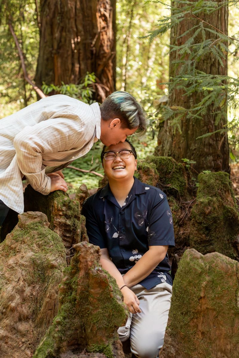 Couple at the Henry Cowell Redwoods State Park one person is kissing the forehead of the other who is wearing glasses, a blue button up and slacks and smiling and sitting on a rock 