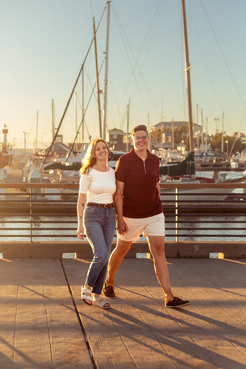 Couple walking and holding hands at Jack London Square in front of boats one is wearing a white shirt, a belt, and blue jeans and the other is wearing a red shirt and white shorts 