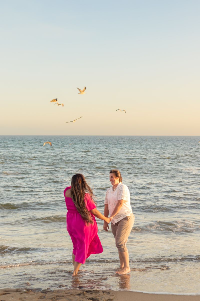 Couple holding hands in front of the ocean while seagulls fly overhead one is wearing a neon pink dress and the other is wearing a white shirt and tan pants they are both laughing with each other 