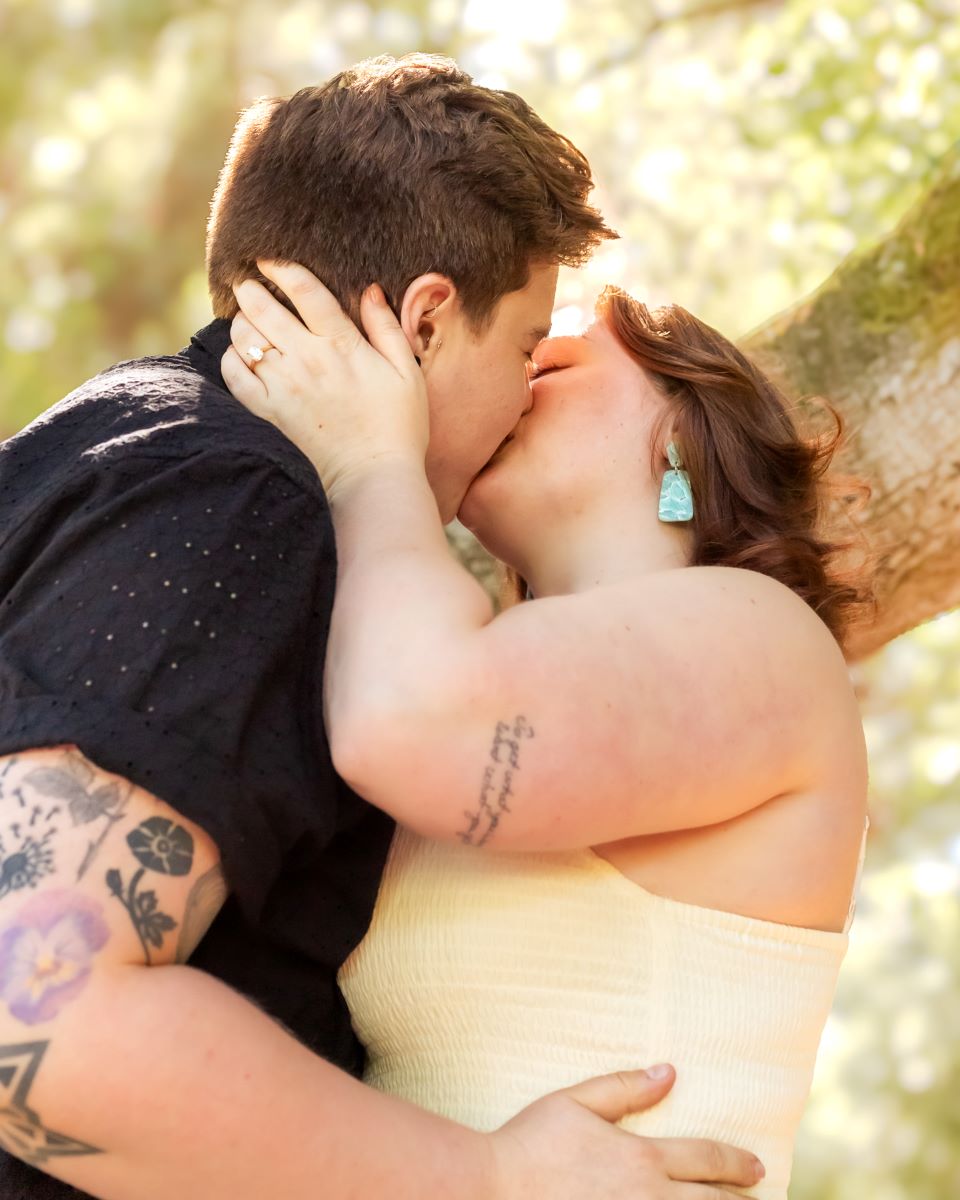 couple kissing each other in front of a tree branch - one of them is wearing a yellow dress and blue earrings and the other is wearing a black shirt - you can see the engagement ring of the one who is wearing the yellow dress and blue earrings 