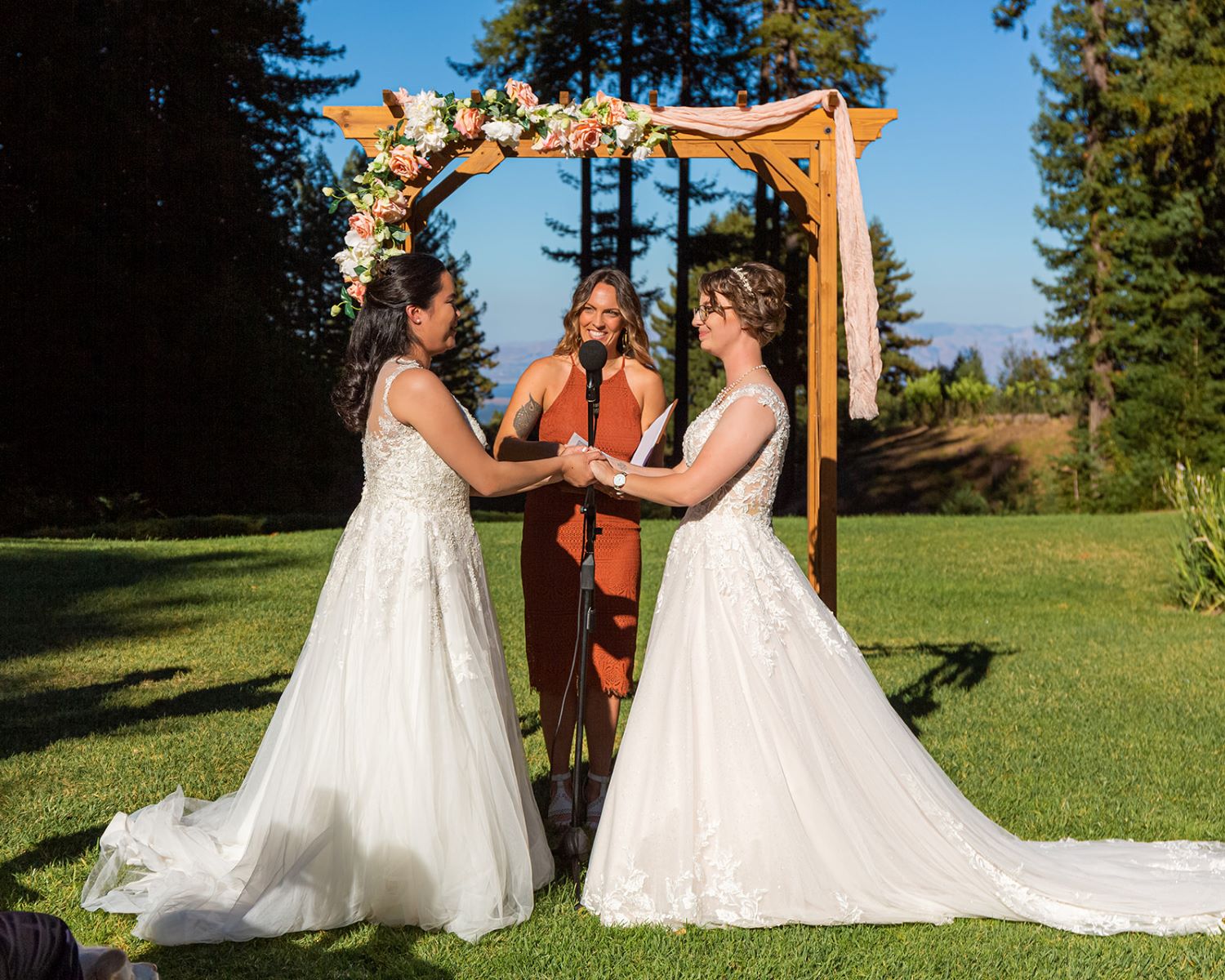 A couple at their wedding ceremony with their officiant in front of a wood arch and both are wearing white wedding dresses