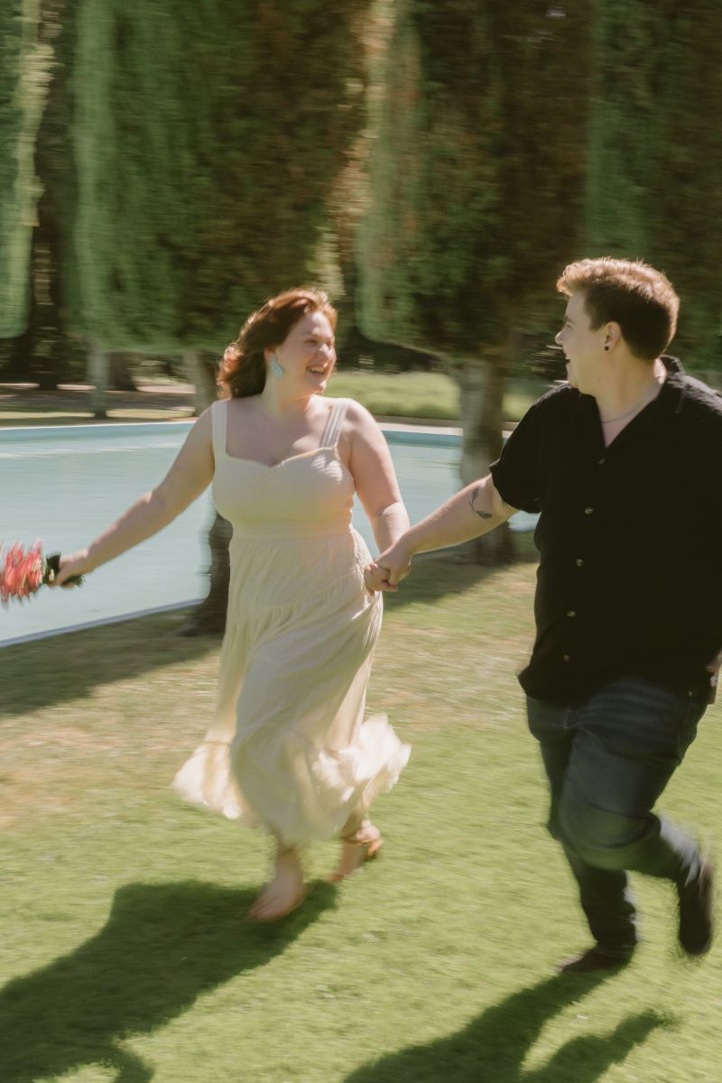 A couple running together and laughing next to a reflecting pool one is weawring a cream colored dress and holding flowers in one hand the other is wearing a black shirt and jeans