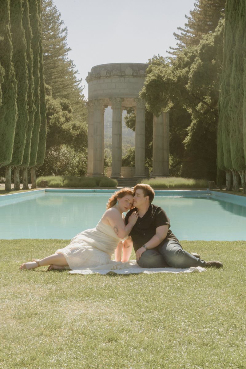 A couple sitting on the grass in front of a reflecting pool and one is wearing a cream colored dress and brown heels the other is wearing a black shirt and jeans and they are both leaning towards each other 