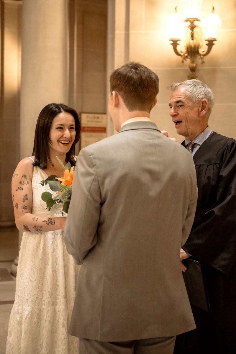 A couple at their wedding ceremony inside of a city hall one is wearing a white dress and holding flowers the other is wearing a gray suit 