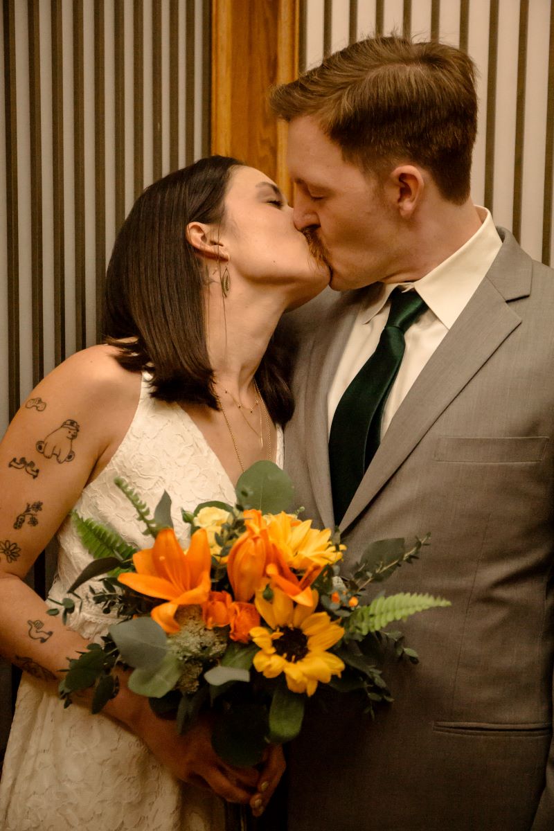 A couple kissing each other one is wearing a white wedding dress and has tattos on their arm and is holding a bouquet of flowers and the other is wearing a gray tuxedo and a green tie 