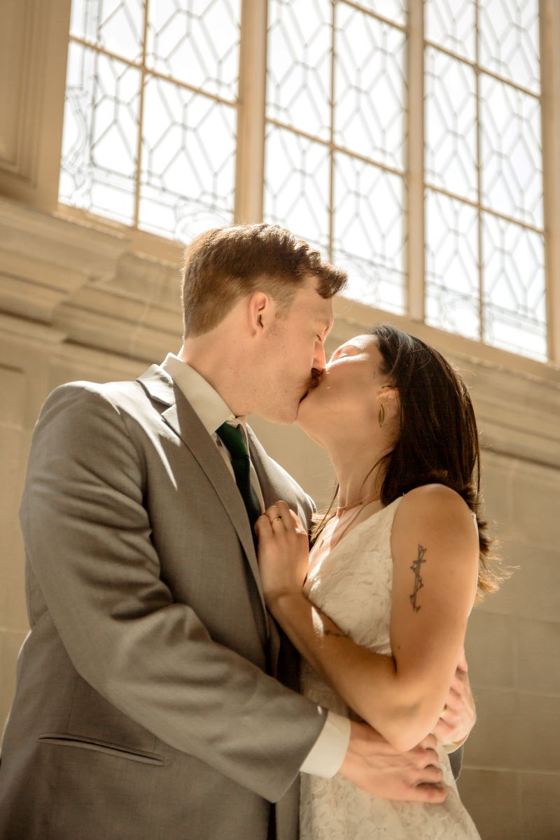 A couple kissing in front of a window one is wearing a white dress and the other is weraing a gray tuxedo and a green tie 