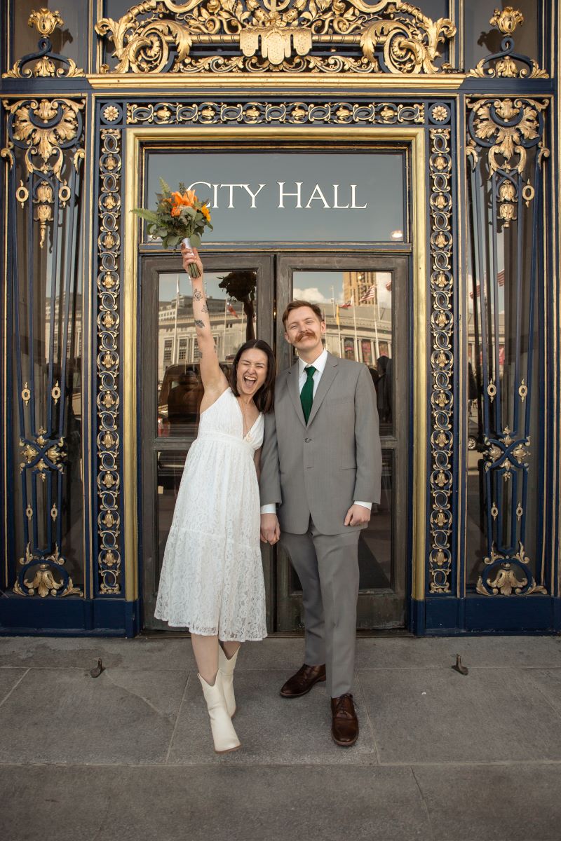 A couple celebrating in front of a city hall building one is wearing a white wedding dress, has tattos, and is throwing up their arm and holding a bouquet of flowers and the other is wearing a gray tuxedo and they are holding hands 