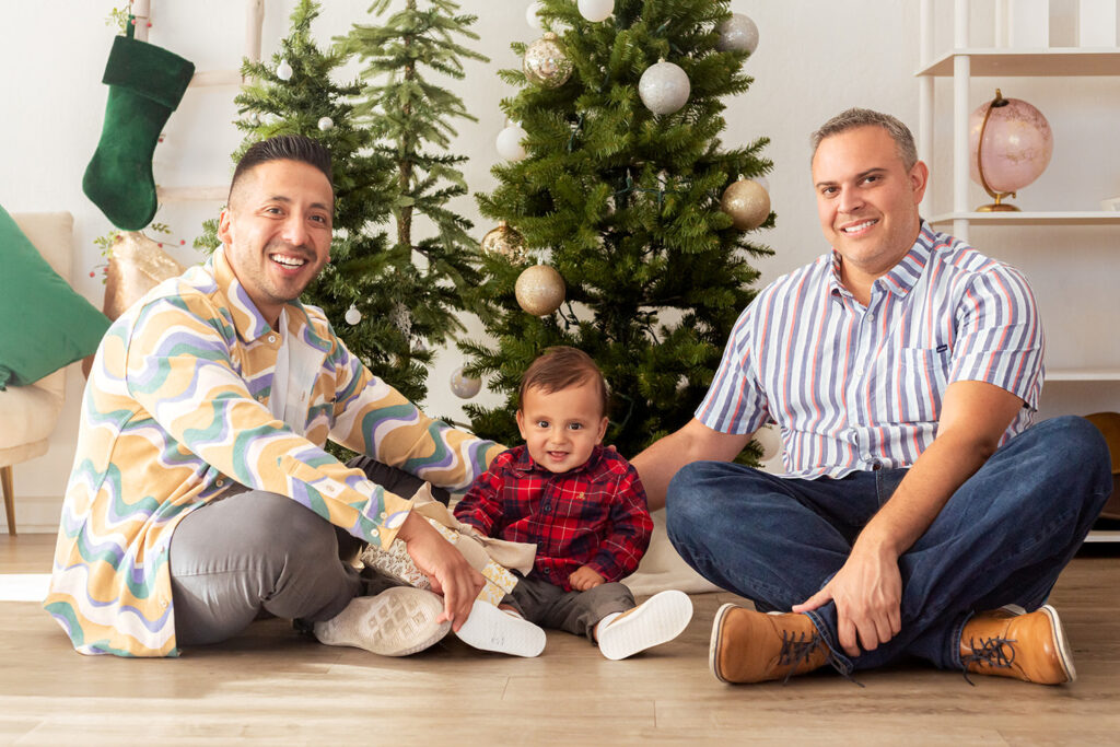 A couple sits on the ground with their child in front of Christmas trees.