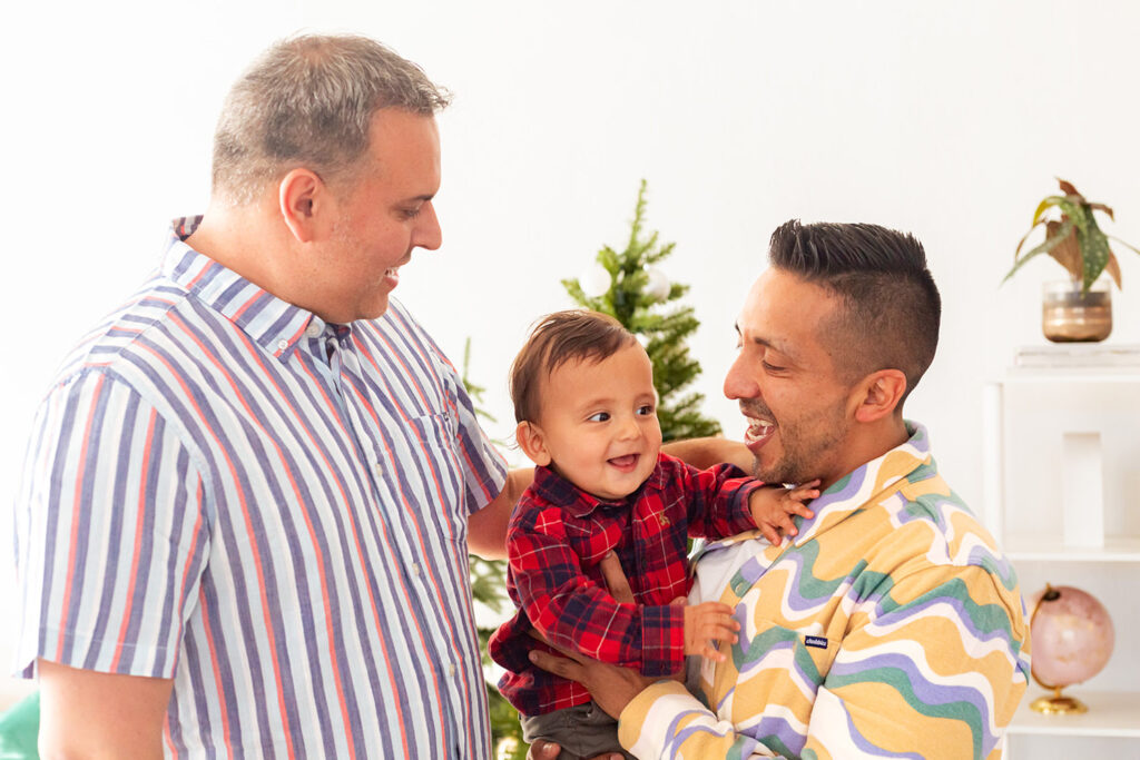 A family of three laughs and smiles. A Christmas tree can be seen behind them.