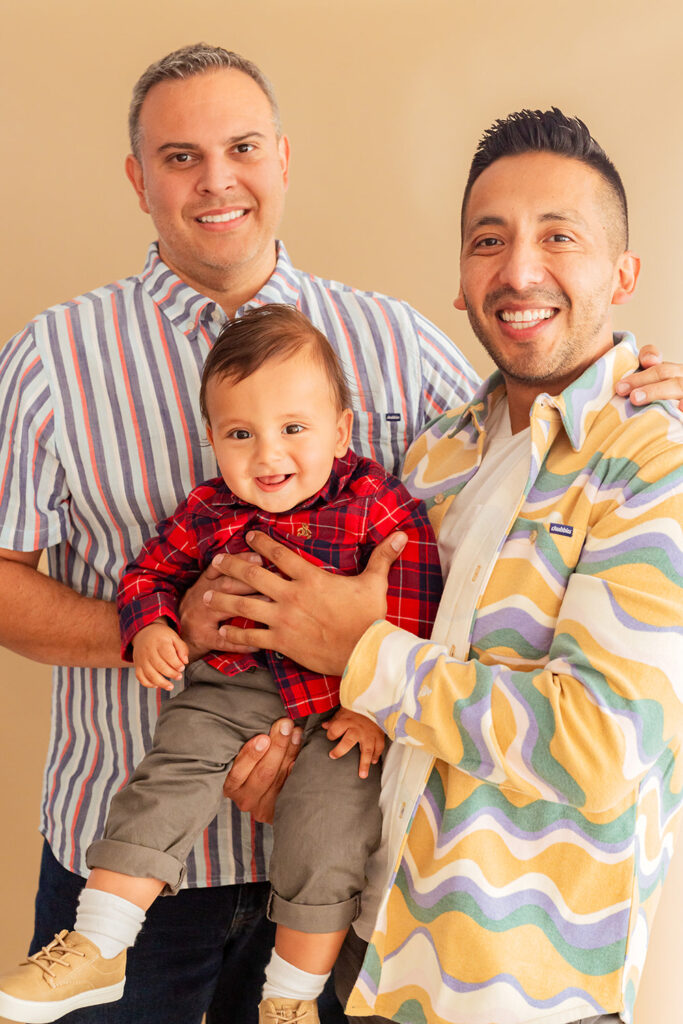 A family of three smiles in front of a tan background.