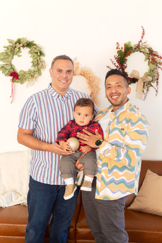 A couple holds their child who holds a golden ornament. Three wreaths are on the wall behind them.