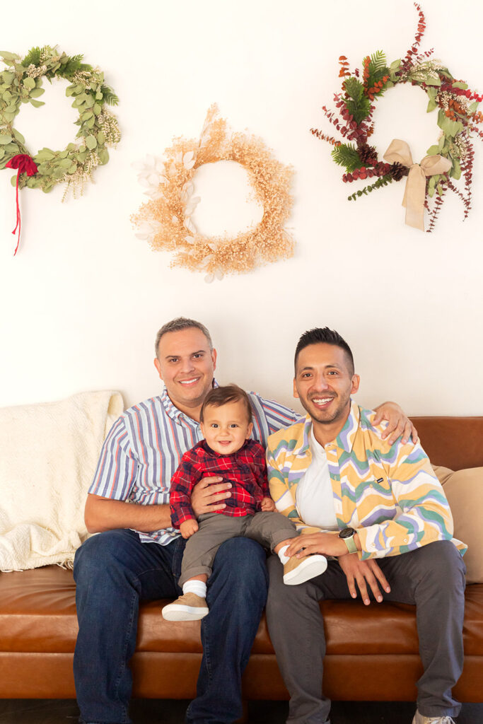 Two parents sit on a leather couch smiling while holding their child.