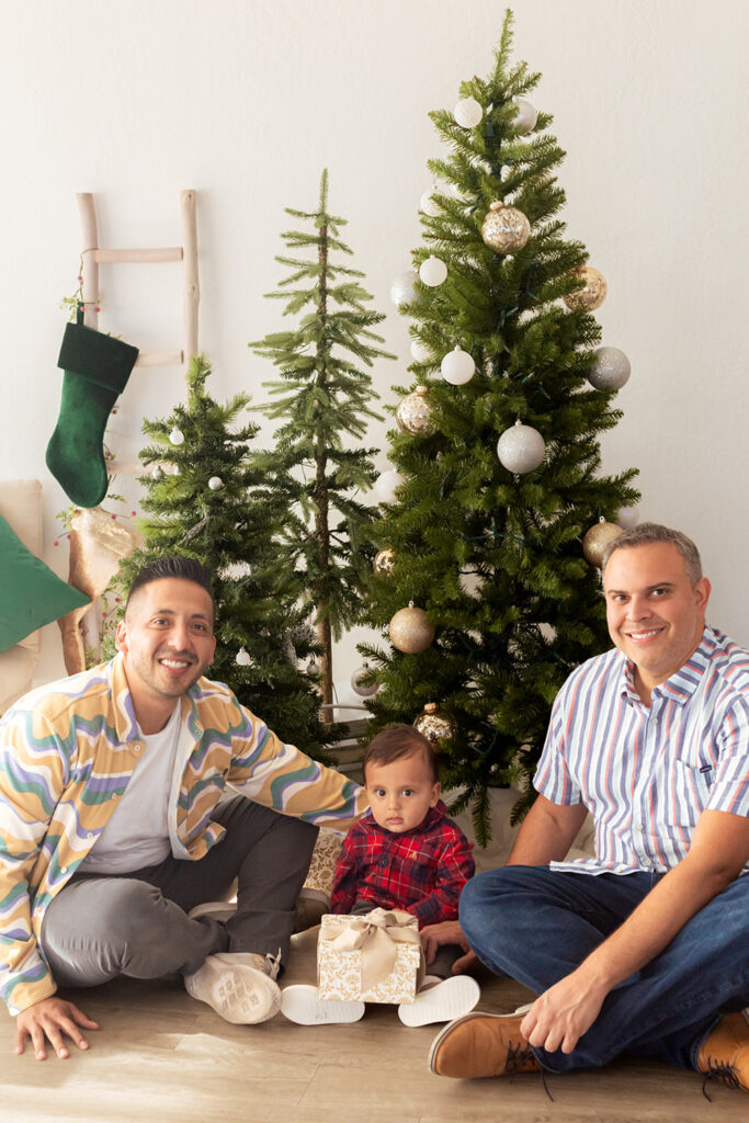 Two parents sit in front of Christmas trees with their child who holds a present.