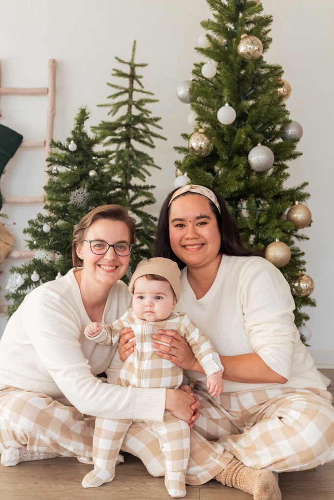 A family of three sits on the floor with three Christmas trees behind them.