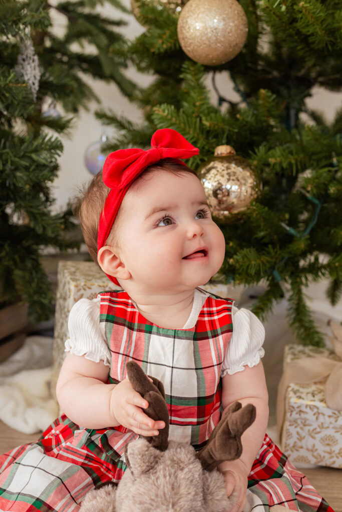 A child in a red plaid dress smiles in front of a tree.