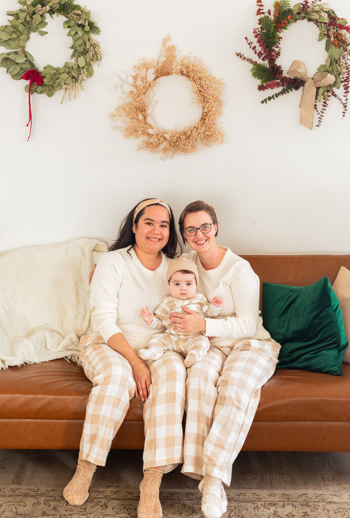 A couple sits on a leather couch holding their child with wreaths behind them.