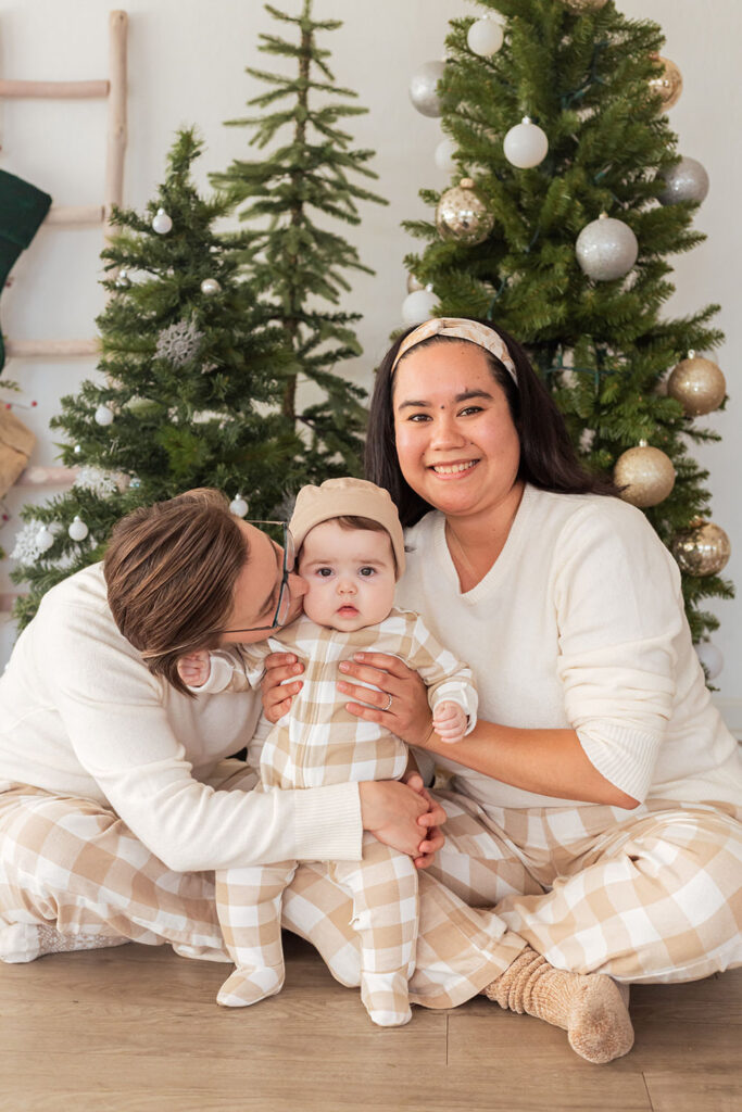 Two parents sit on the ground in matching pajamas while one kisses their child on the cheek.