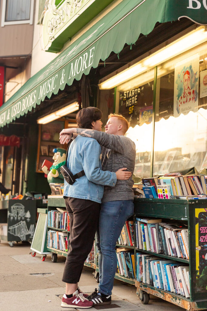 A couple with their arms around each other leaning on a cart of books outside a book store 