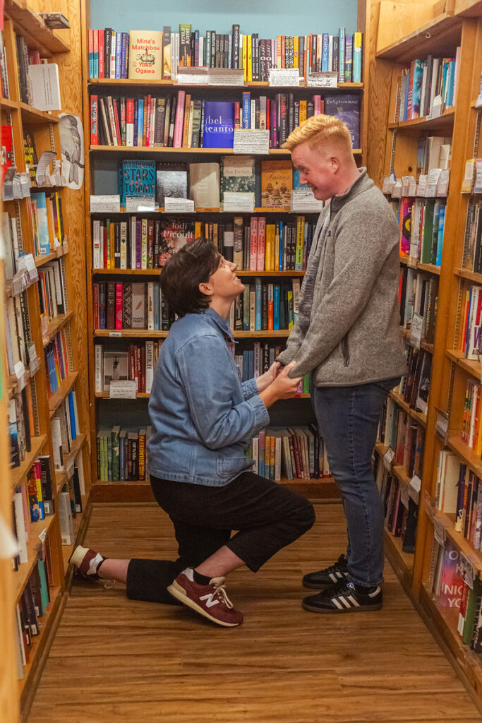 A person smiling as their partner is on one knee proposing in a bookstore 