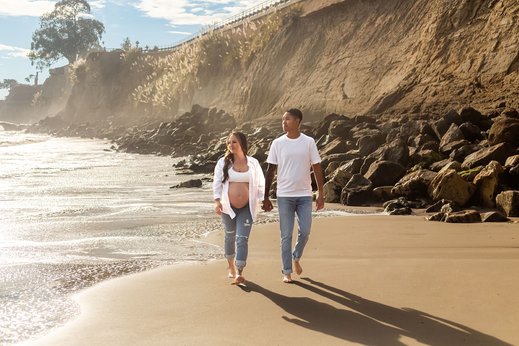 A couple walking hand in hand along the beach next to the ocean with rocks and cliffs behind them one is wearing a white t-shirt and jeans and the other is pregnant and weraing a white bra, white button up shirt and jeans both are barefoot and looking towards the ocean