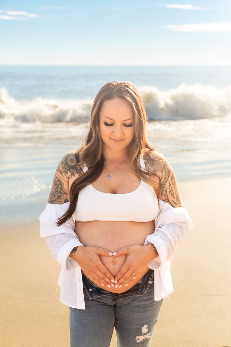 A pregnant woman is making a heart shape with her hands and holding her hands against her belly she has painted white nails and tattoos on her shoulders and is wearing a white button up shirt and whtie bra and jeans and she is looking down towards her stomach she is standing on the sand with the ocean behind her 