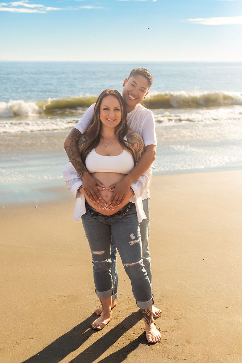 A couple standing in the sand next to the ocean one has their arms wrapped around their partner and both are smiling and they are both wearing white shirts and blue jeans and one is pregnant and they both have their hands on her belly 