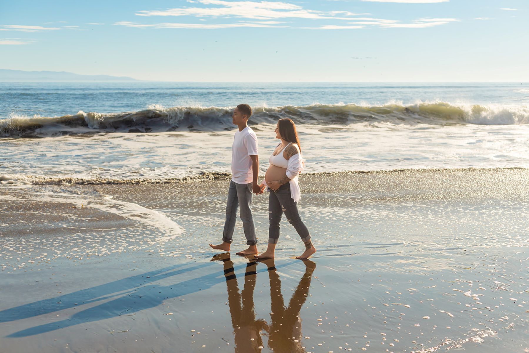 A couple walking hand in hand on the beach with the ocean behind them one is wearing a white shirt and blue jeans and the other is a pregnant woman who is wearing a white bra and blue jeans and white button up and they are both barefoot