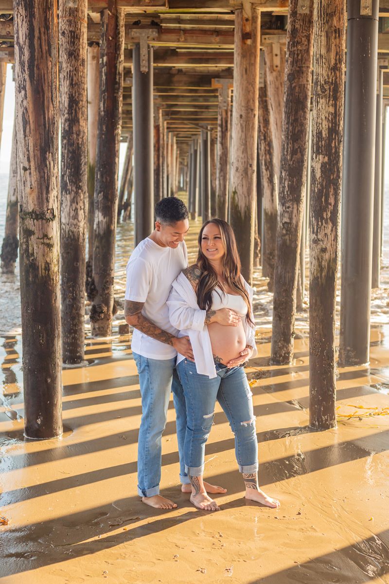 A couple standing underneath a wooden pier in the sand barefoot laughing with each other one has their hands on their partner's hips and is wearing a white shirt and blue jeans the the other is a pregnant woman who is wearing a white bra, white button up and blue jeans 