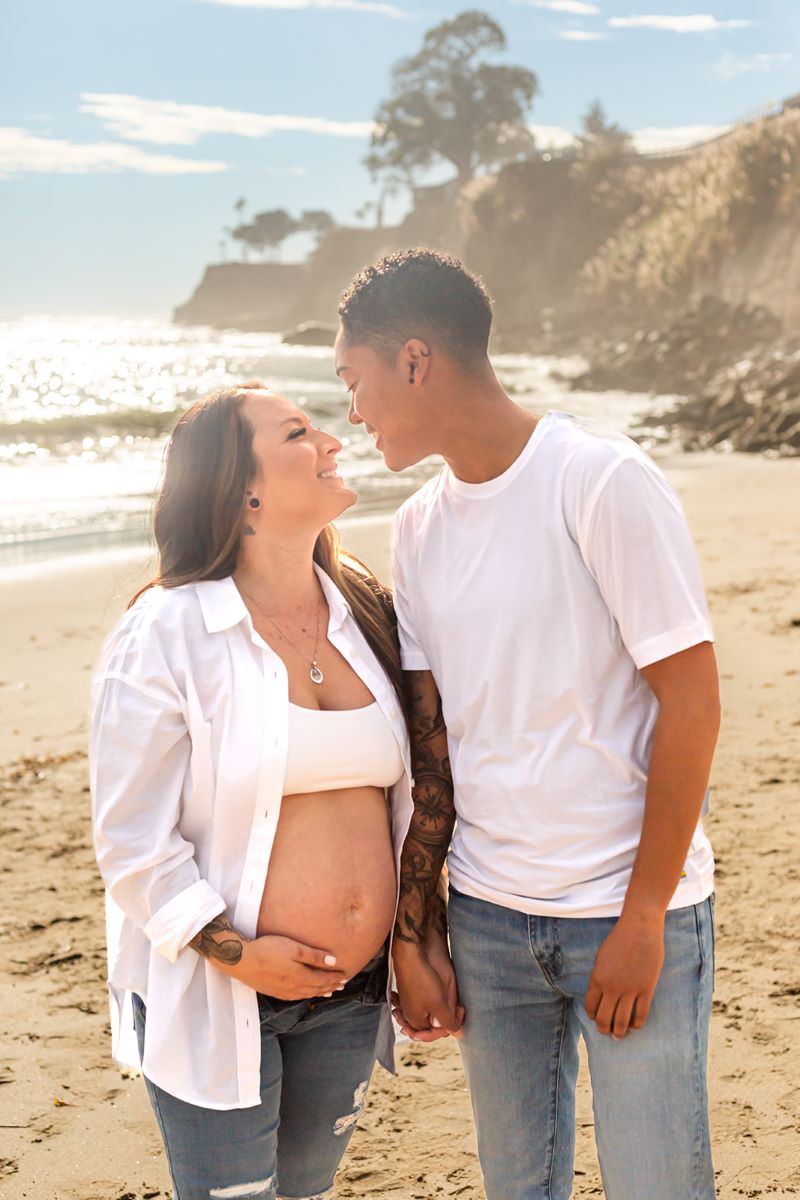 A couple together in the sand ont he beach looking at each other and smiling one is wearing a white shirt and blue jeans and the other is a pregnant woman who is wearing a white bra and blue jeans and white button up shirt and the are both holding hands with each other