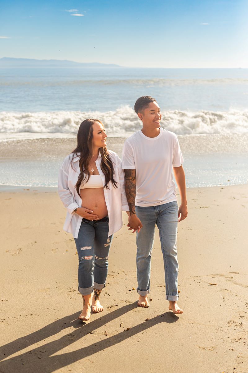 A couple walking hand in hand on the beach with the ocean behind them one is wearing a white shirt and blue jeans and they are both barefoot and the other is a pregnant woman wearing a white button up, white bra, and blue jeans