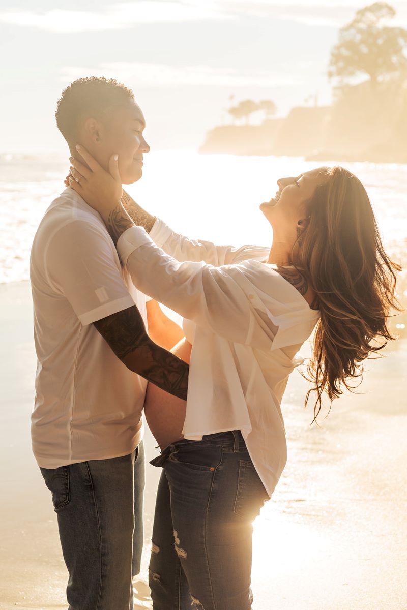 A couple together on the beach with their arms wrapped around each other and smiling one is wearing a white shirt and jeans the other is leaning slightly backwards and she is pregnant and wearing a white button up shirt and jeans