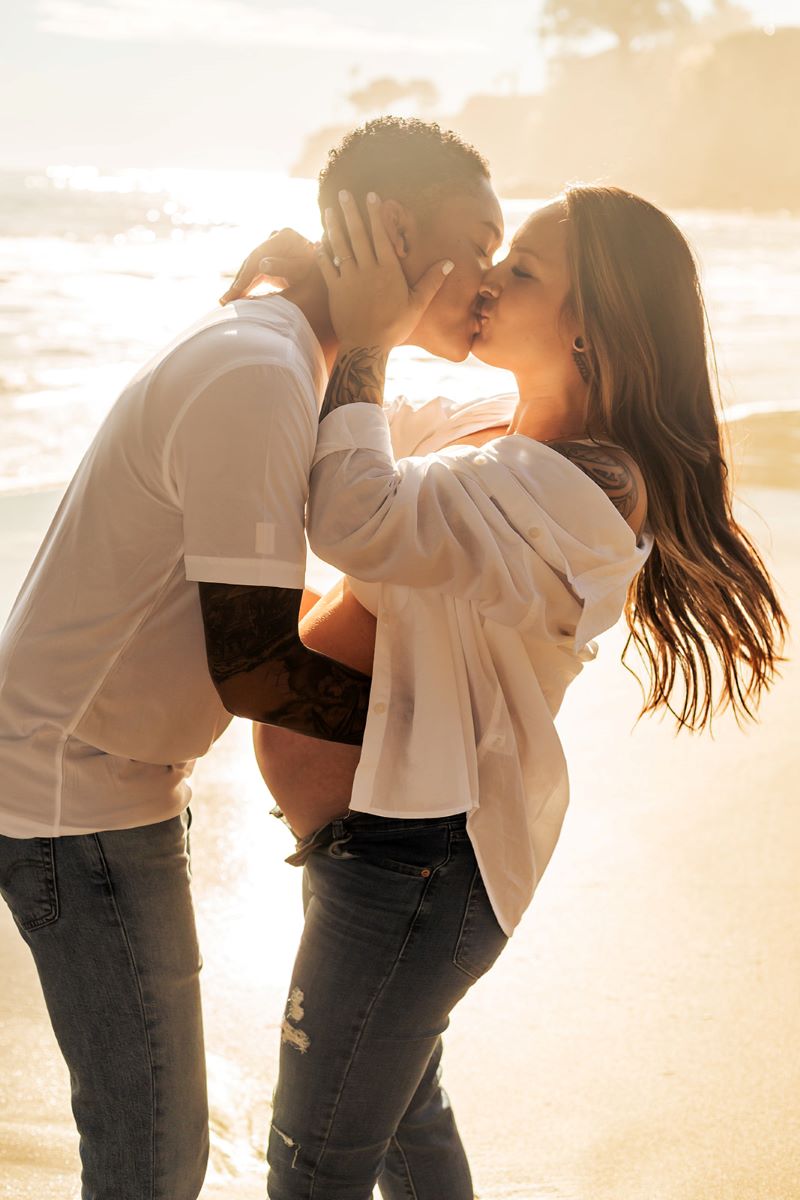 A couple on the beach kissing each other one is a pregnant woman wearing a white button up and jeans and she has her hands on her partner's face who is wearing a white shirt and jeans 