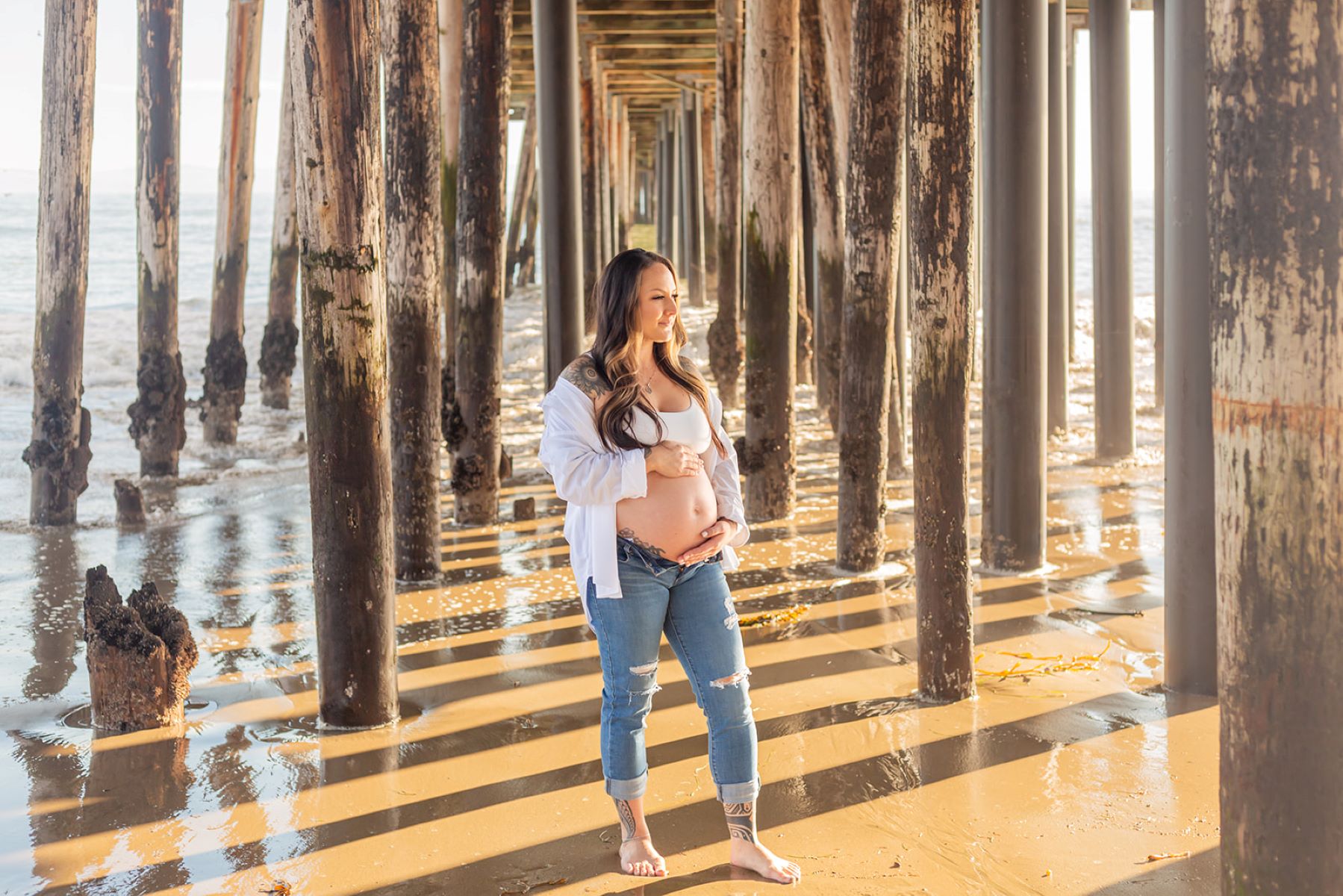 A pregnant woman standing underneath a wooden pier in the water barefoot and she is wearing a white bra and blue jeans and a white button up 