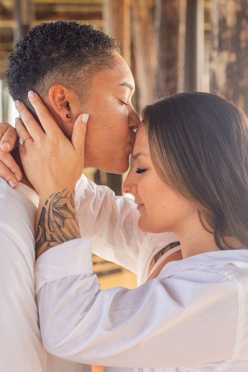 A couple standing underneath a wooden pier one is kissing their partner on the forehead and the other is leaning in for the kiss