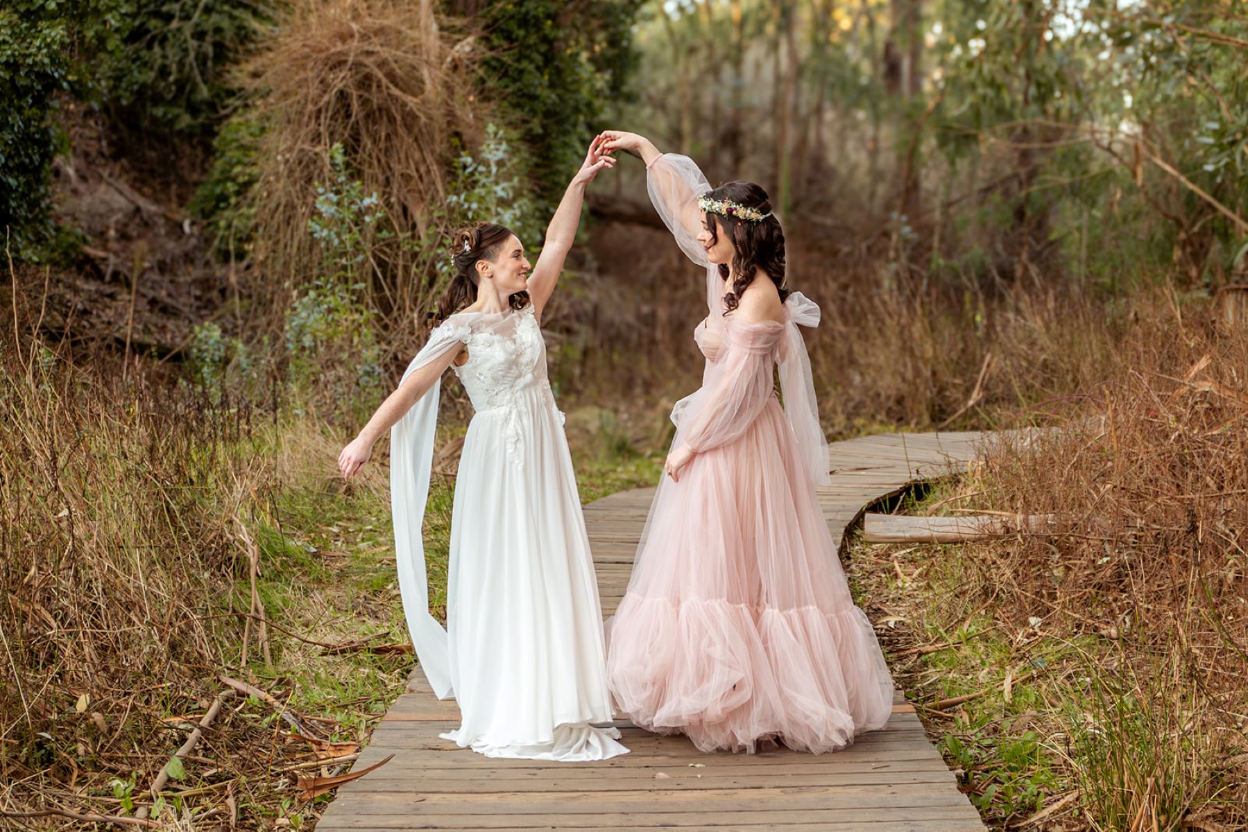 A couple dancing on a wooden path in a forest and one is wearing a white gown and the other is wearing a pink gown the partner is the pink gown is spinning their partner in the white gown