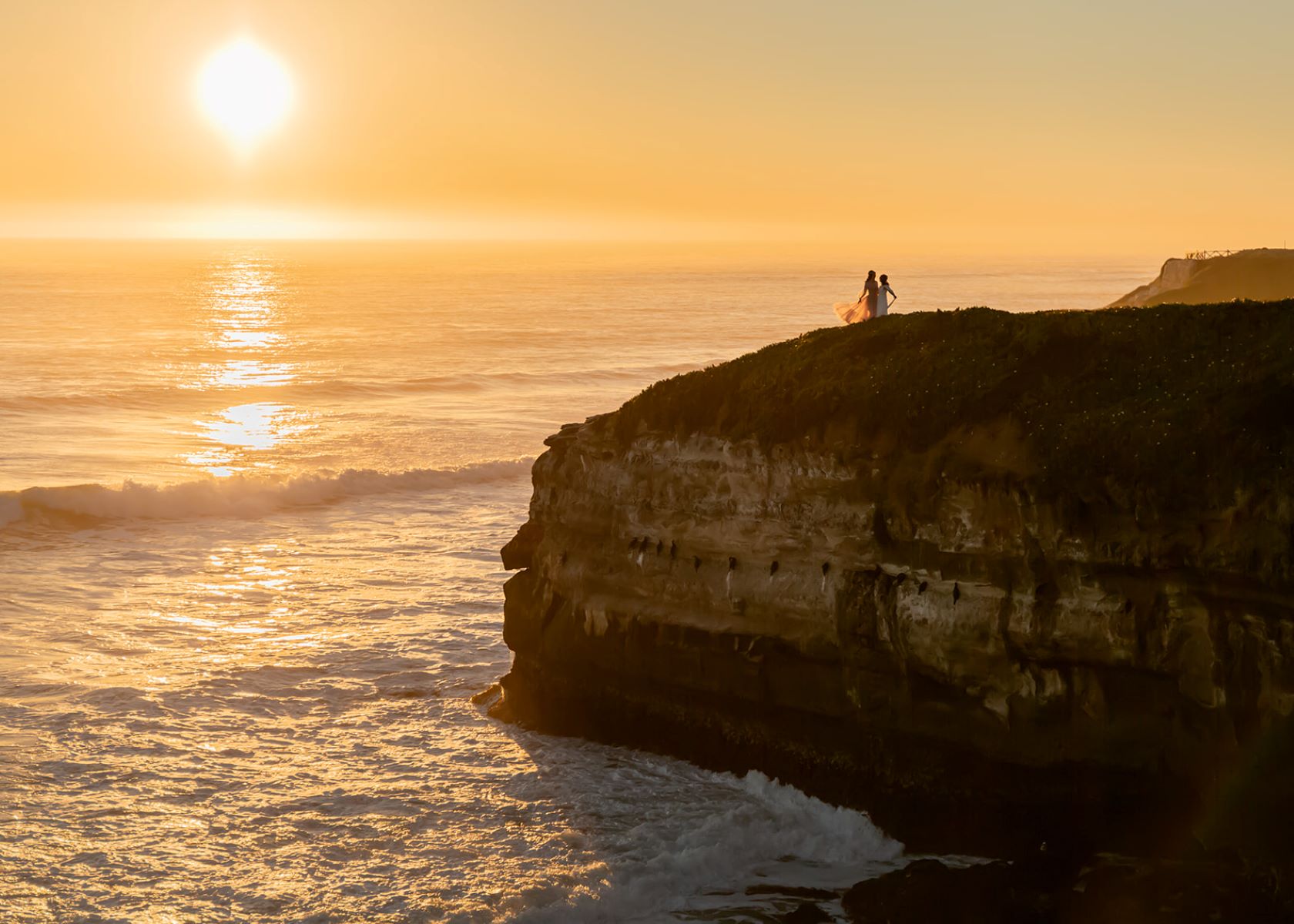 A couple on a cliffside at sunset next to the ocean one is wearing a white gown the other is wearing a pink gown