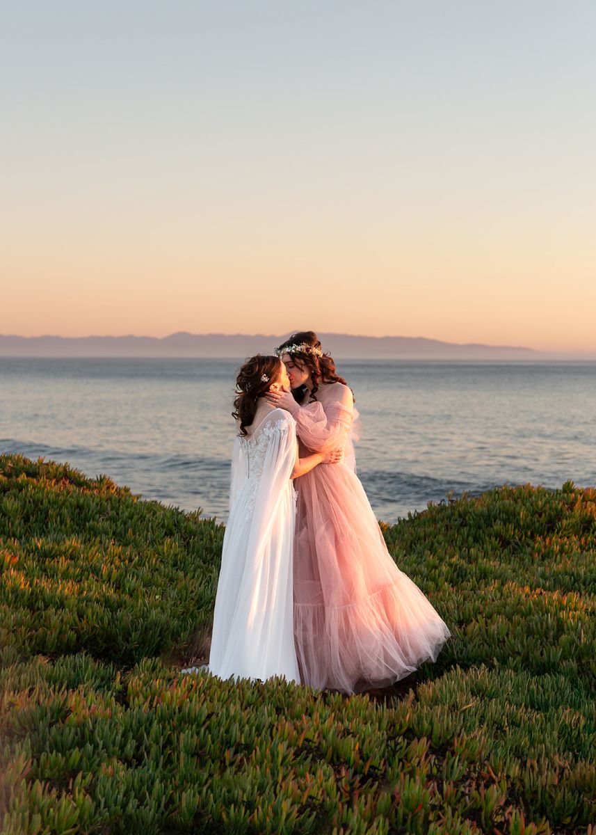 A couple kissing at sunset in the grass on a cliffside with the ocean behind them one is wearing a pink gown and the other is wearing a white gown